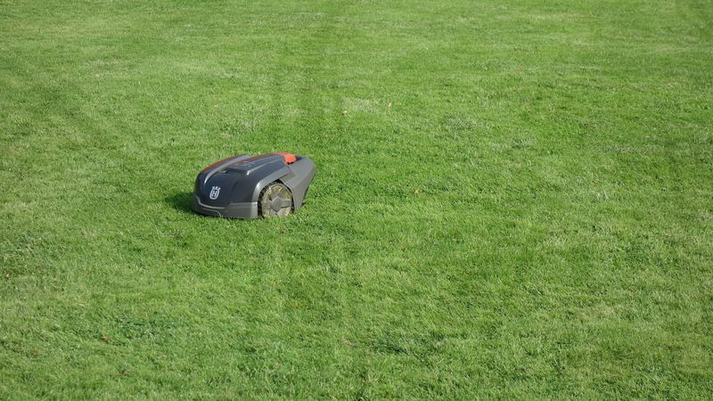 A small grey Husqvarna robot mower sitting on a large, freshly striped green lawn