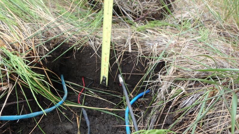 Soil sensor cables running into a freshly dug hole in grassy ground, with a tape measure showing the depth