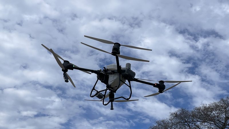 A large black agricultural spray drone with four rotors flying against a cloudy blue sky